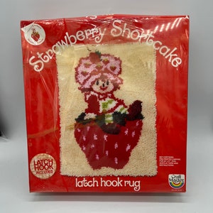 Puede incluir: Una caja roja y blanca con un kit de alfombra de gancho de cierre con Strawberry Shortcake. La caja incluye lienzo preimpreso, hilo acr&iacute;lico e instrucciones. La imagen muestra a Strawberry Shortcake sentada en una fresa roja.