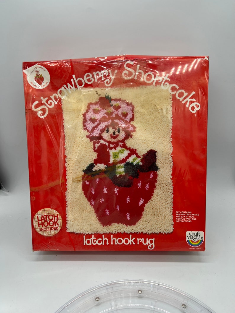 Puede incluir: Una caja roja y blanca con un kit de alfombra de gancho de cierre con Strawberry Shortcake. La caja incluye lienzo preimpreso, hilo acr&iacute;lico e instrucciones. La imagen muestra a Strawberry Shortcake sentada en una fresa roja.