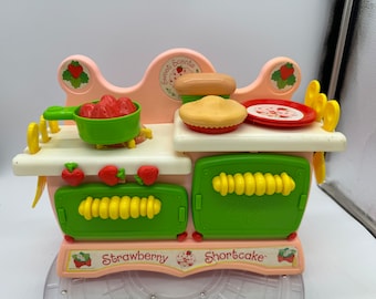 May include: A pink plastic toy kitchen playset with a Strawberry Shortcake theme. The kitchen has two ovens, a stovetop, and a counter with a pie, a loaf of bread, and a bowl of strawberries. The kitchen is decorated with yellow and green accents.