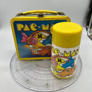 May include: A yellow Pac-Man lunchbox and thermos. The lunchbox has a blue background with Pac-Man and ghosts. The thermos has a white background with Pac-Man and ghosts.