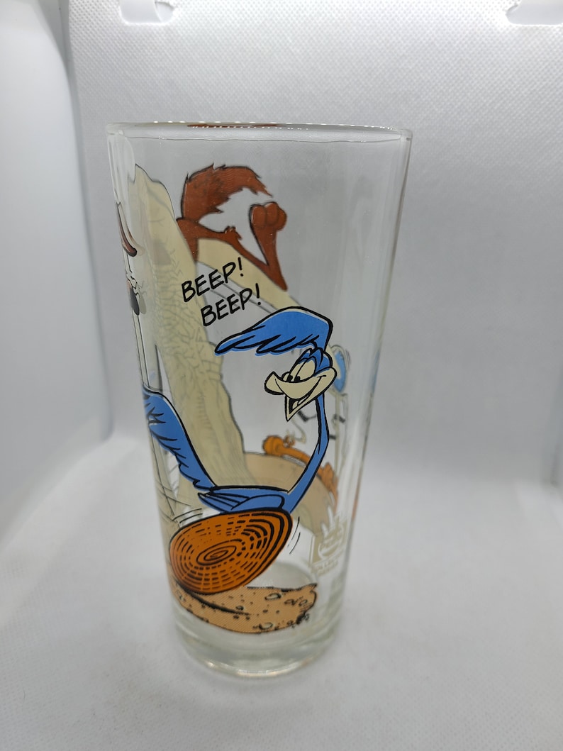 May include: A clear glass featuring a cartoon image of the Road Runner and Wile E. Coyote. The Road Runner is blue and running with a large orange disc. The text "Beep! Beep!" is printed on the glass.