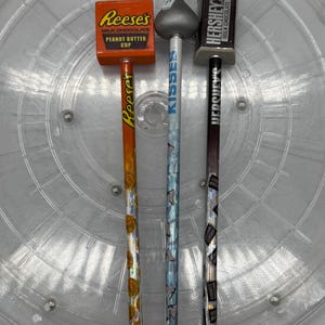May include: Three decorative pencils topped with candy-themed toppers. The left pencil is orange with Reese's branding, the centre pencil is light blue with a Hershey's Kisses topper, and the right pencil is dark brown with Hershey's branding.