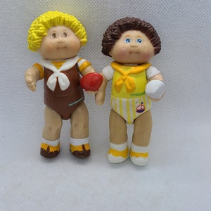 May include: Two Cabbage Patch Kids dolls wearing different outfits. The doll on the left is wearing a brown and yellow outfit with a red apple. The doll on the right is wearing a yellow and white outfit with a white ball.