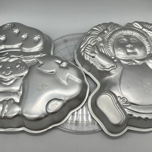 Puede incluir: Dos moldes para pasteles de metal vintage, uno con forma de Strawberry Shortcake y el otro con forma de muñeca Cabbage Patch Kid. Ambos moldes son de color plateado.