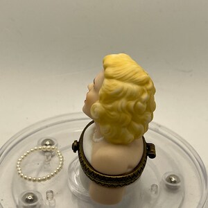 May include: A porcelain bust of a woman with blonde hair, wearing a white dress and a pearl necklace. The bust is a hinged box with a gold-toned metal frame.