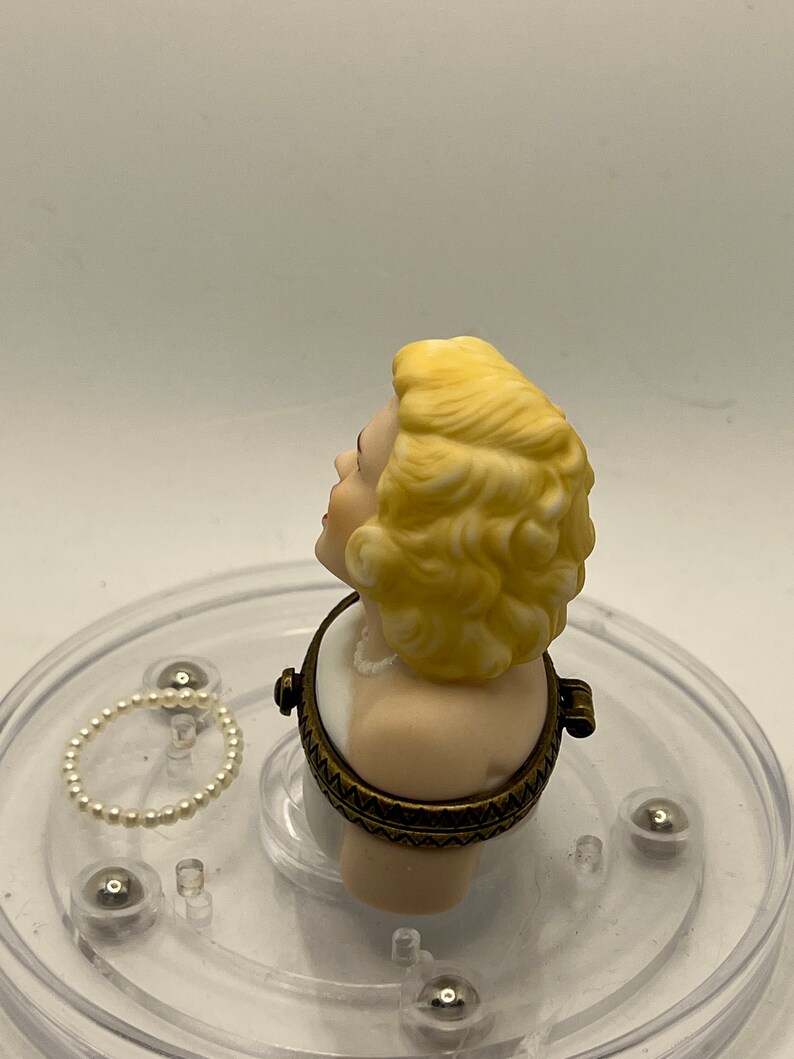 May include: A porcelain bust of a woman with blonde hair, wearing a white dress and a pearl necklace. The bust is a hinged box with a gold-toned metal frame.