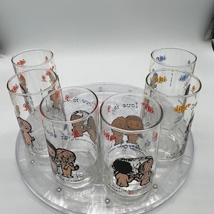 May include: Set of six clear glass tumblers with cartoon illustrations of Peanuts characters. Each glass features a different Peanuts character and a quote about love. The glasses are arranged on a clear plastic rotating tray.