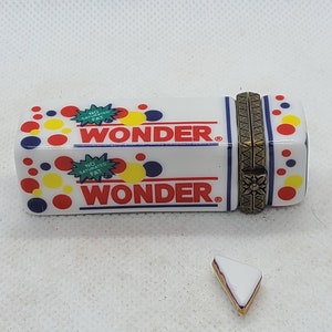 Può includere: Una scatola di ceramica bianca con un design Wonder Bread. La scatola ha pois rossi, gialli e blu e le parole "No Saturated Fat" e "Wonder" sul davanti. La scatola è aperta e un piccolo panino triangolare di ceramica bianca è seduto davanti alla scatola.
