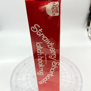 Puede incluir: Una caja roja con el texto "Strawberry Shortcake latch hook rug" y "Craft Master" en ella. La caja est&aacute; sobre un marco de alfombra de gancho de pl&aacute;stico transparente.