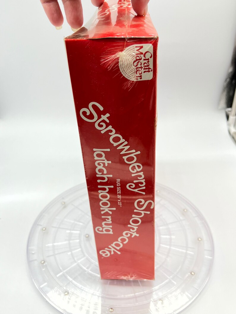 Puede incluir: Una caja roja con el texto "Strawberry Shortcake latch hook rug" y "Craft Master" en ella. La caja est&aacute; sobre un marco de alfombra de gancho de pl&aacute;stico transparente.