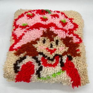 Op de afbeelding: Een vintage naaldviltkussen met een cartoonfiguur met rood haar, een roze hoed en een rood-witte jurk. Het kussen is gemaakt van een pluche, textuurrijke stof.