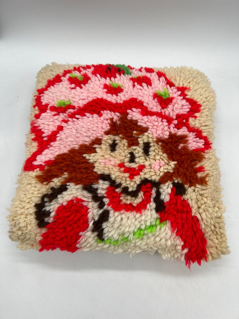 Op de afbeelding: Een vintage naaldviltkussen met een cartoonfiguur met rood haar, een roze hoed en een rood-witte jurk. Het kussen is gemaakt van een pluche, textuurrijke stof.