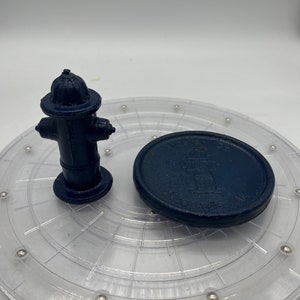 以下が含まれることがあります： 透明なプラスチック製のターンテーブルの上に、濃い青色の消火栓と、濃い青色の円形のディスクがあり、消火栓のデザインが描かれています。