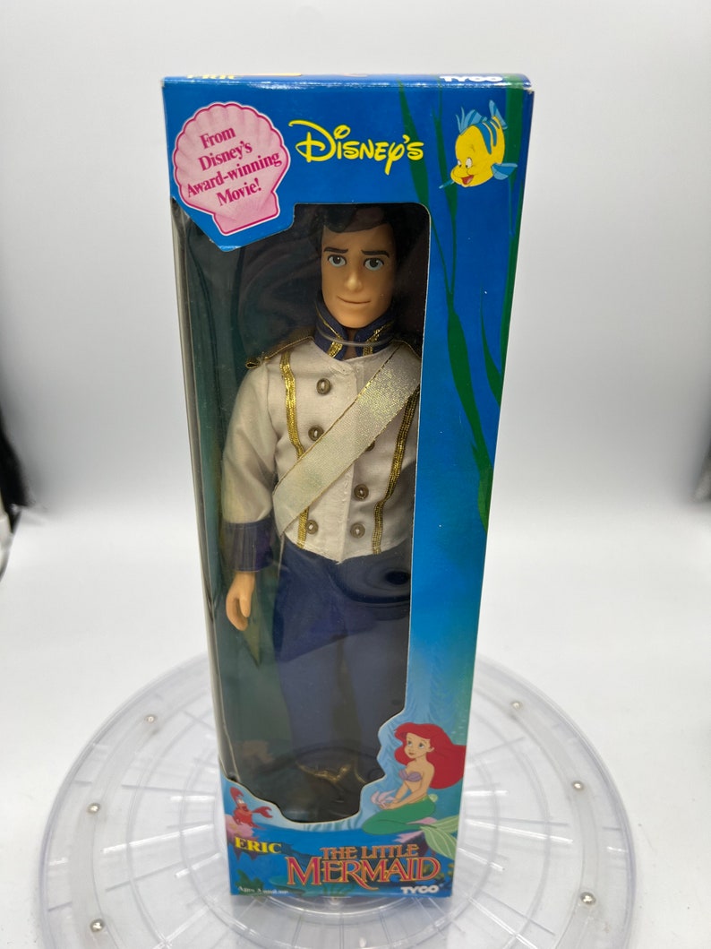 Pode incluir: Uma boneca de pl&aacute;stico do Pr&iacute;ncipe Eric do filme A Pequena Sereia da Disney. A boneca est&aacute; vestida com um uniforme branco e azul com detalhes dourados. A boneca est&aacute; embalada em uma caixa azul com uma imagem de Ariel e Linguado na frente.