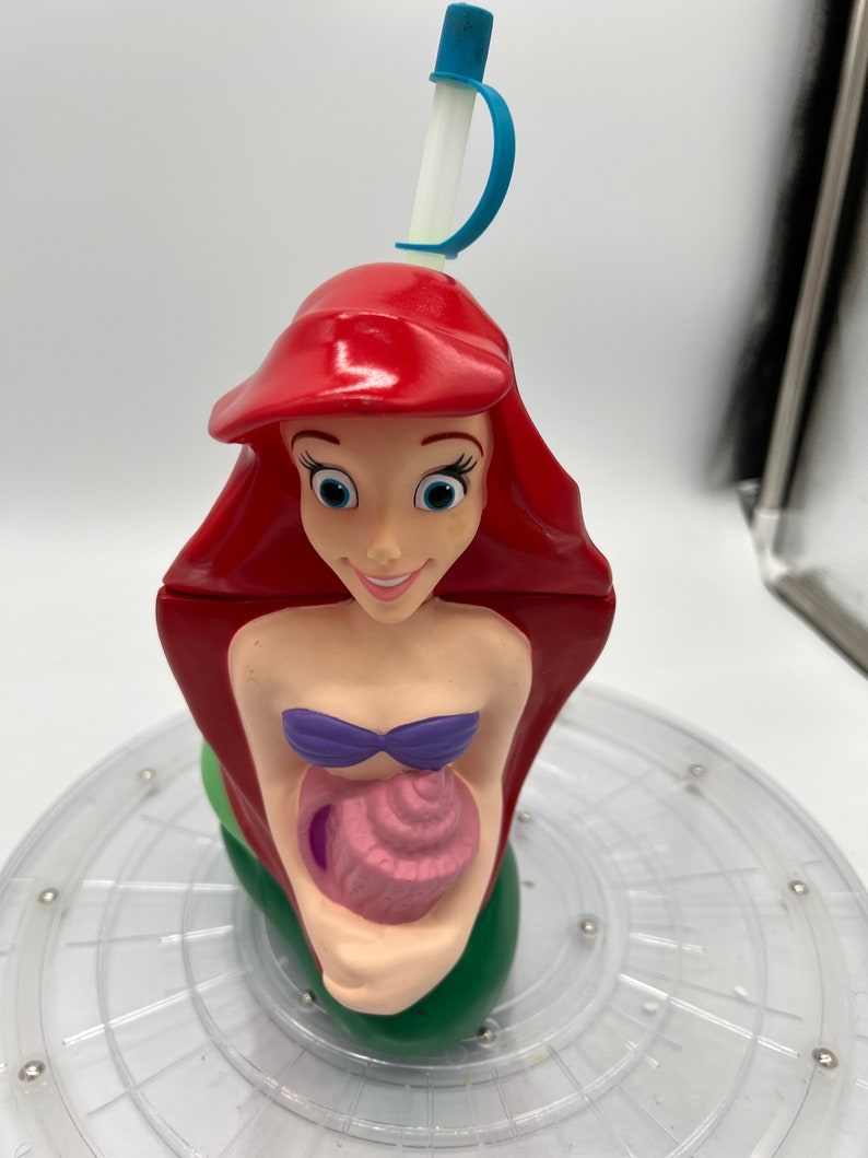 Op de afbeelding: Een rode, groene en paarse plastic beker in de vorm van Ariel, de Kleine Zeemeermin, die een roze schelp vasthoudt. De beker heeft een blauwe en witte rietje.