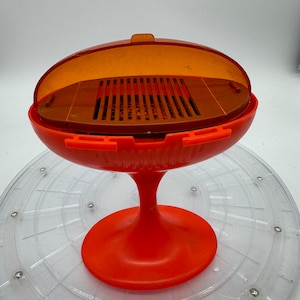 Könnte beinhalten: Ein orangefarbenes und braunes Kunststoff-Barbecue-Spielzeug im Retro-Stil. Der Grill hat einen abgerundeten Sockel und einen transparenten orangefarbenen Deckel. Der Innenraum ist braun mit einem Grillrost. Der Sockel ist leuchtend orange.
