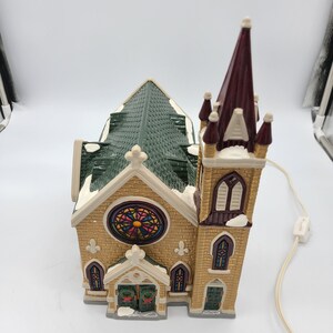 K&ouml;nnte beinhalten: Eine beleuchtete Keramik-Kirchenfigur mit einem gr&uuml;nen Dach, Buntglasfenstern und einem hohen Turm. Die Kirche ist mit wei&szlig;en Schneeakzenten verziert und hat ein Lichtschnur.