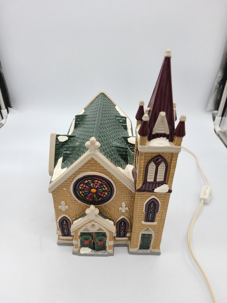 K&ouml;nnte beinhalten: Eine beleuchtete Keramik-Kirchenfigur mit einem gr&uuml;nen Dach, Buntglasfenstern und einem hohen Turm. Die Kirche ist mit wei&szlig;en Schneeakzenten verziert und hat ein Lichtschnur.