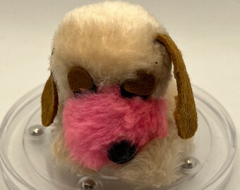 Può includere: Un piccolo cane di peluche bianco e soffice con orecchie marroni e un naso rosa. Il cane è seduto su una base di plastica trasparente con accenti in metallo argentato.