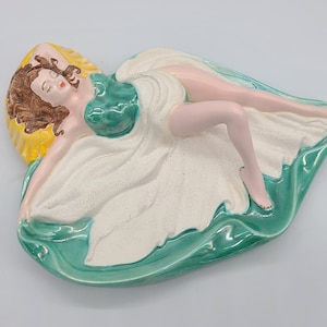 Puede incluir: Una figurita de cerámica de una mujer recostada sobre una superficie ondulada verde y blanca. La mujer lleva un vestido verde y blanco y tiene el pelo castaño.