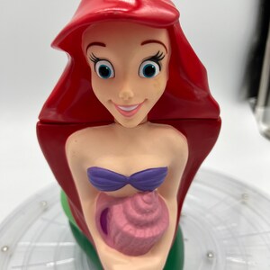 Op de afbeelding: Een rode en groene plastic beker in de vorm van Ariel, de Kleine Zeemeermin, die een roze schelp vasthoudt. De beker heeft een rietjegat en is klaar voor een verfrissend drankje.