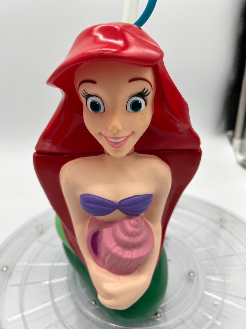 Op de afbeelding: Een rode en groene plastic beker in de vorm van Ariel, de Kleine Zeemeermin, die een roze schelp vasthoudt. De beker heeft een rietjegat en is klaar voor een verfrissend drankje.