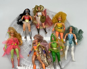 Op de afbeelding: Set van zeven vintage action figuren, met vrouwelijke personages met kleurrijke kostuums en accessoires. De figuren staan op een transparante plastic displaystandaard.