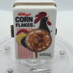 May include: A white box with a red rooster and the words "Kellogg's Corn Flakes" printed on it. The box is a miniature replica of a cereal box and has a small spoon attached to it. The box is sitting on a clear plastic base with silver balls around the edge.
