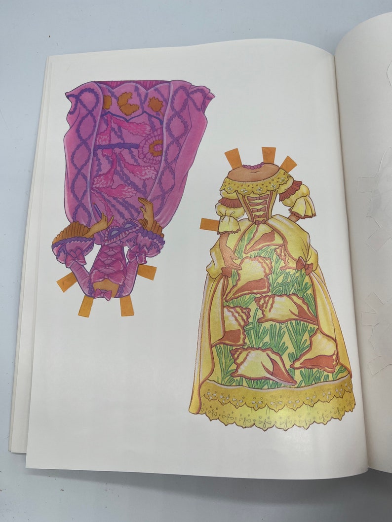 Puede incluir: Dos mu&ntilde;ecas de papel con atuendos diferentes. La primera mu&ntilde;eca lleva un vestido rosa y morado con un borde de encaje blanco. La segunda mu&ntilde;eca lleva un vestido amarillo con un patr&oacute;n de conchas marinas y hojas verdes.