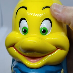 Puede incluir: Figurita de pl&aacute;stico amarilla de un pez de dibujos animados con una bufanda azul. El pez tiene ojos verdes, una boca roja y una sonrisa blanca.