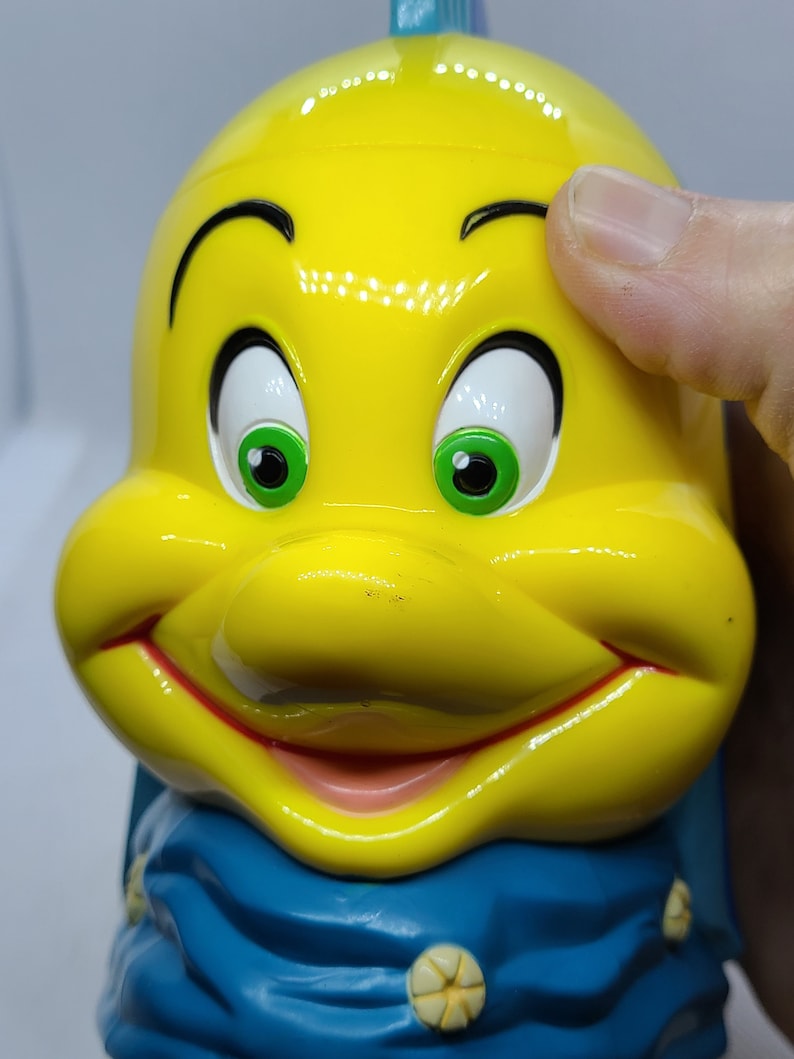Puede incluir: Figurita de pl&aacute;stico amarilla de un pez de dibujos animados con una bufanda azul. El pez tiene ojos verdes, una boca roja y una sonrisa blanca.
