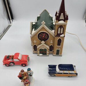 K&ouml;nnte beinhalten: Ein Keramik-Weihnachtsdorf-Set mit einer Kirche mit einem Buntglasfenster, einem roten Pickup-Truck, einem blauen Kombi und einer Familienfigur.