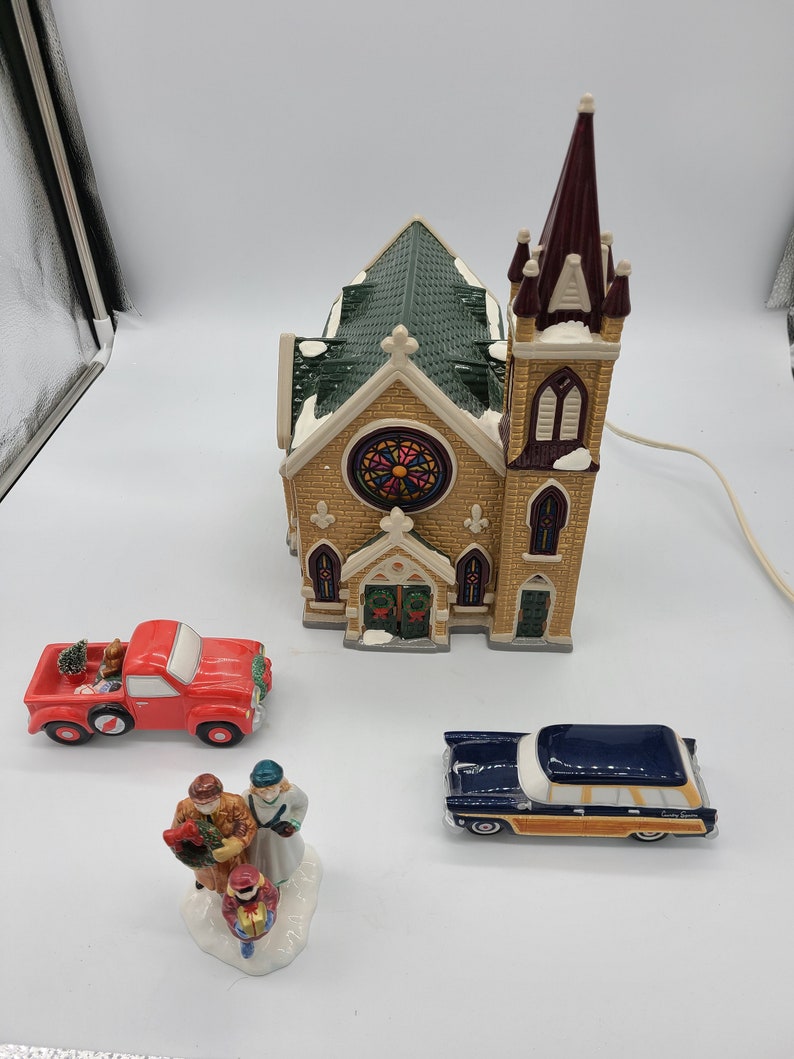 K&ouml;nnte beinhalten: Ein Keramik-Weihnachtsdorf-Set mit einer Kirche mit einem Buntglasfenster, einem roten Pickup-Truck, einem blauen Kombi und einer Familienfigur.