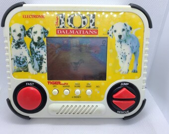 Op de afbeelding: Een wit-zwart handheld elektronisch spel met het thema 101 Dalmatiërs van Disney. Het spel heeft een scherm dat een cartoon Dalmatiër puppy laat zien die rent. Het spel heeft knoppen met de labels "Fast", "Jump", "Rescue", "Off", "Sound", "Max Score", "On/Start", en "ACL".