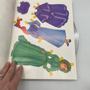 Puede incluir: Un libro de mu&ntilde;ecas de papel con una mujer pelirroja con un vestido verde, un vestido azul y una capa morada. El libro incluye varios accesorios de mu&ntilde;ecas de papel, como un sombrero y un lazo.