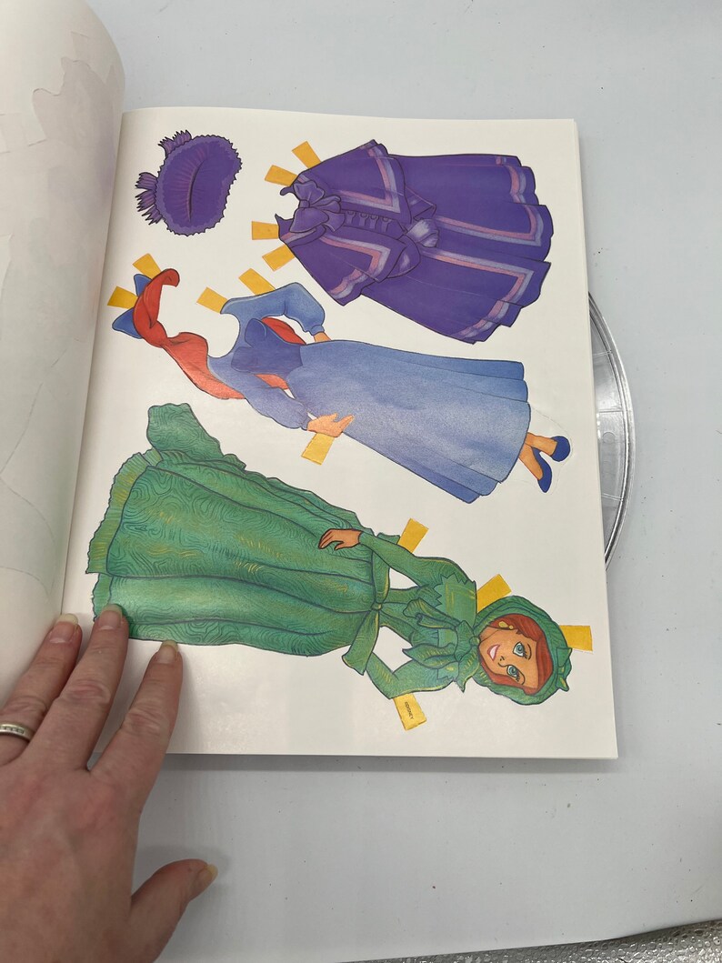 Puede incluir: Un libro de mu&ntilde;ecas de papel con una mujer pelirroja con un vestido verde, un vestido azul y una capa morada. El libro incluye varios accesorios de mu&ntilde;ecas de papel, como un sombrero y un lazo.