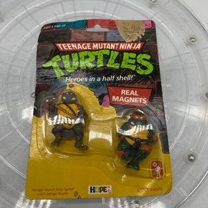 May include: Two Teenage Mutant Ninja Turtles action figures with real magnets. The figures are packaged on a yellow card with the words "Teenage Mutant Ninja Turtles" and "Heroes in a half shell!" The card also says "Real Magnets" and "Stock #60215".