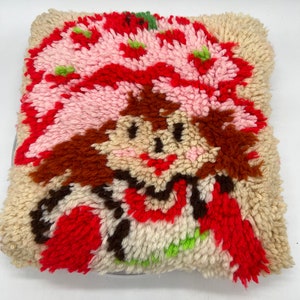 Op de afbeelding: Een vintage naaldviltkussen met een cartoonfiguur met rood haar, een roze hoed en een rode jurk. Het figuur glimlacht en heeft bruine ogen. Het kussen is gemaakt van een zachte, pluche stof en is in goede staat.
