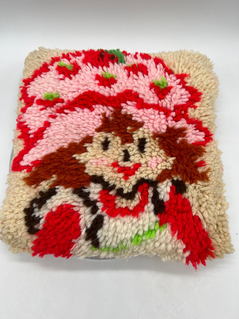 Op de afbeelding: Een vintage naaldviltkussen met een cartoonfiguur met rood haar, een roze hoed en een rode jurk. Het figuur glimlacht en heeft bruine ogen. Het kussen is gemaakt van een zachte, pluche stof en is in goede staat.