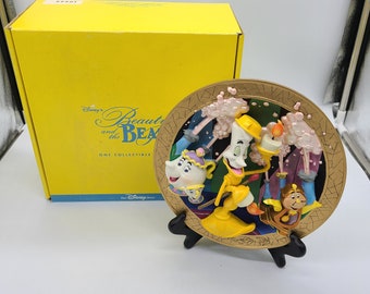 Könnte beinhalten: Ein goldfarbener Keramikteller mit einem 3D-Design von Figuren aus Disneys Die Schöne und das Biest. Die Figuren sind Lumière, Herr von der Tanne, Mrs. Potts und Chip. Der Teller steht auf einem schwarzen Ständer und trägt die Aufschrift "Disney's Beauty and the Beast" und "One Collectible".