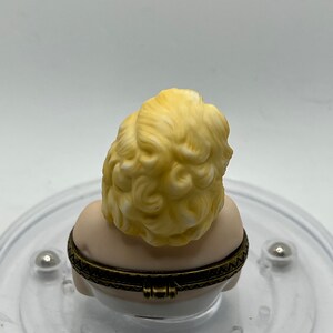 May include: A porcelain trinket box featuring a bust of a woman with blonde hair. The box is closed and has a gold-colored hinge.