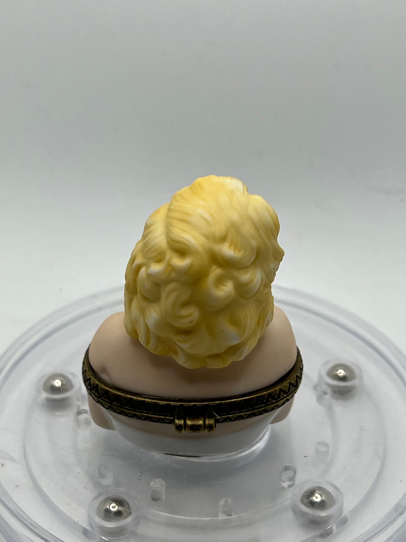 May include: A porcelain trinket box featuring a bust of a woman with blonde hair. The box is closed and has a gold-colored hinge.