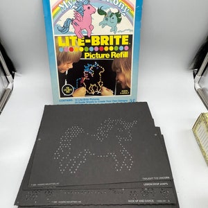 May include: A Lite-Brite picture refill set featuring My Little Pony characters. The box shows a pink pony with a rainbow mane and a blue pony with a purple mane. The set includes 12 Lite-Brite pictures and 24 guide sheets to create your own designs.