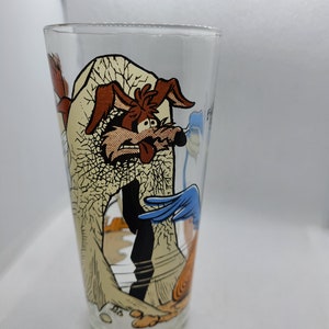 May include: A glass drinking glass featuring a cartoon image of Wile E. Coyote and Road Runner. The glass depicts a scene of Wile E. Coyote falling from a cliff with Road Runner running away.