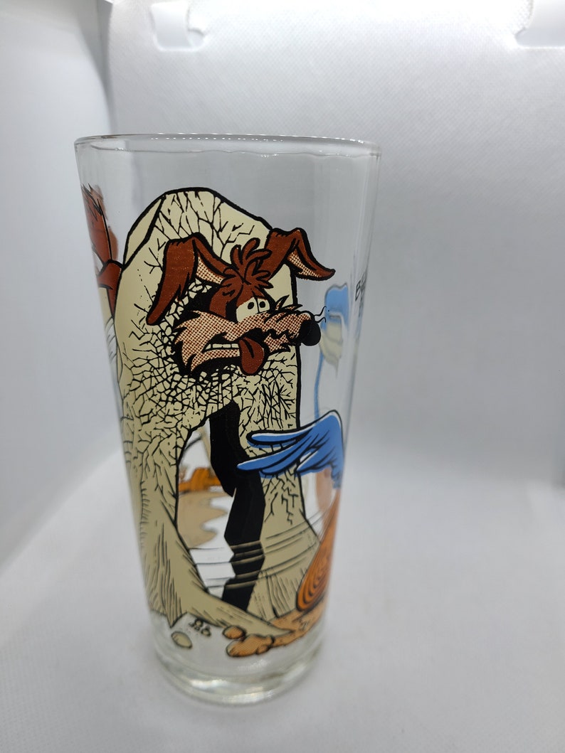 May include: A glass drinking glass featuring a cartoon image of Wile E. Coyote and Road Runner. The glass depicts a scene of Wile E. Coyote falling from a cliff with Road Runner running away.