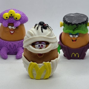 May include: Three Halloween-themed McDonald's Happy Meal toys. A purple octopus with three eyes, a mummy with a spider on its head, and a Frankenstein with a green face and purple clothing.