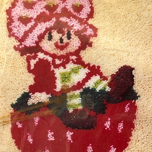 Puede incluir: Una fresa bordada de color rojo y blanco con una ni&ntilde;a sonriente que lleva un sombrero rosa y blanco sentada encima. La ni&ntilde;a sostiene un objeto a rayas verde y blanco.