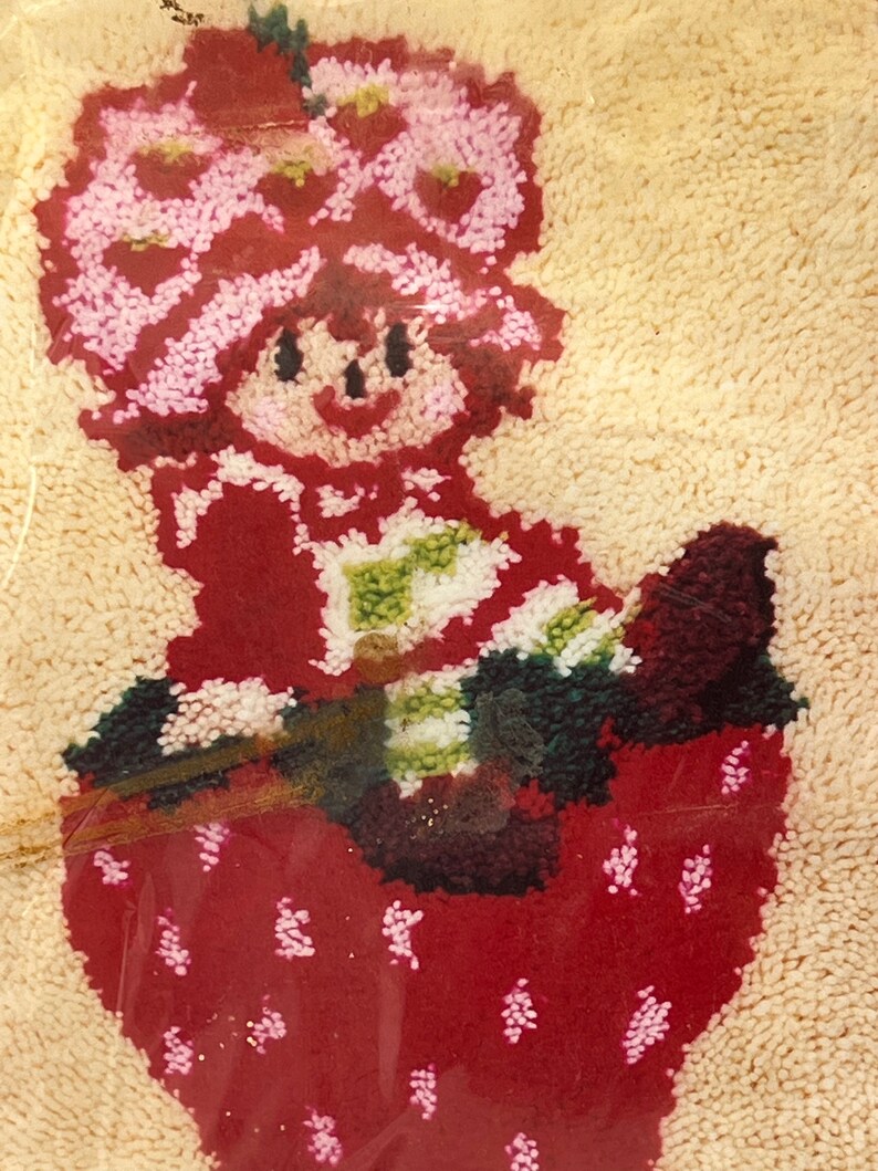 Puede incluir: Una fresa bordada de color rojo y blanco con una ni&ntilde;a sonriente que lleva un sombrero rosa y blanco sentada encima. La ni&ntilde;a sostiene un objeto a rayas verde y blanco.