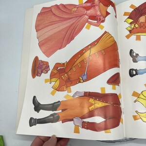Puede incluir: Un conjunto de mu&ntilde;ecas de papel que incluye una princesa con un vestido rosa y un pr&iacute;ncipe con un traje rojo y dorado. El conjunto incluye varias piezas para cada personaje, lo que permite diferentes combinaciones de atuendos.