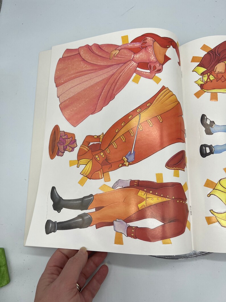 Puede incluir: Un conjunto de mu&ntilde;ecas de papel que incluye una princesa con un vestido rosa y un pr&iacute;ncipe con un traje rojo y dorado. El conjunto incluye varias piezas para cada personaje, lo que permite diferentes combinaciones de atuendos.